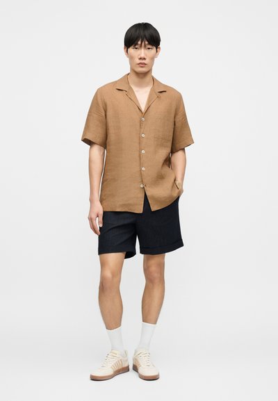 Chemise en lin marron boutonnée avec une poche, associée à un short bleu marine, des chaussettes blanches et des baskets beiges avec des accents marron clair.