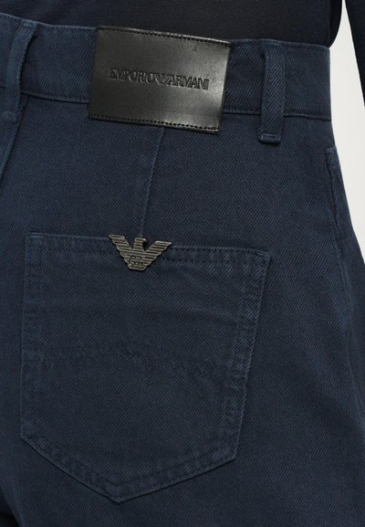 Tumši zila denima bikses ar ādas jostas tagu, uz kura ir iepresēts "EMPORIO ARMANI", papildinātas ar sudrabu ērgļa logotipu uz kabatas.