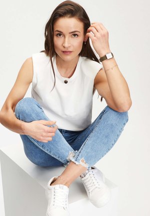 Jonge vrouw die met gekruiste benen op een witte blok zit, gekleed in een witte mouwloze top, gescheurde blauwe jeans, witte sneakers en een horloge.