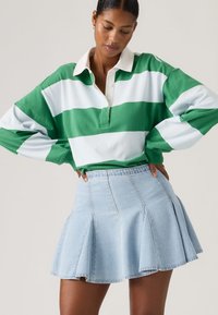 Γυναίκα που  φοράει πράσινο και άσπρο ριγέ μακρυμάνικο πόλο μπλουζάκι, μπαίνετε μέσα σε ένα ανοιχτό μπλε πλισέ μίνι φούστα από τζιν, με τα χέρια στη μέση.