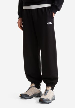 Person iført sorte The North Face joggingbukser med elastiske manchetter og beige vandresko med sorte såler og lynlås.