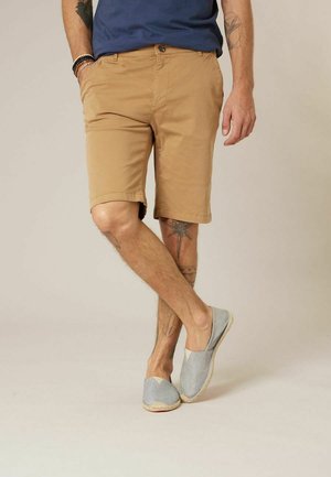 Homme portant un short beige, une chemise bleue, des chaussures grises sans lacets, avec des tatouages sur les jambes et les bras, debout jambes croisées devant un fond uni.