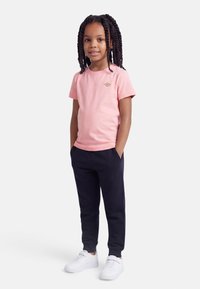 Κορίτσι με μακριά πλεγμένα μαλλιά, ντυμένο με ροζ μπλουζάκι, μαύρο παντελόνι φόρμας και λευκά αθλητικά, στέκεται με τα χέρια στις τσέπες.