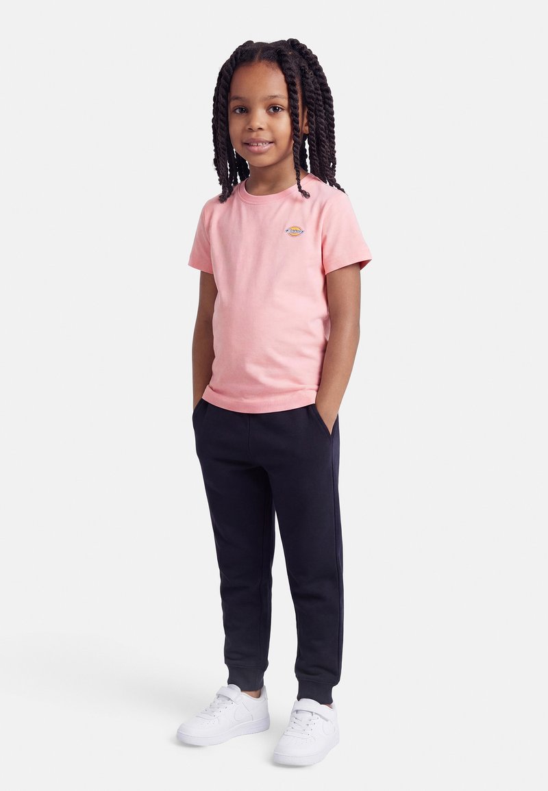 Κορίτσι με μακριά πλεγμένα μαλλιά, ντυμένο με ροζ μπλουζάκι, μαύρο παντελόνι φόρμας και λευκά αθλητικά, στέκεται με τα χέρια στις τσέπες.