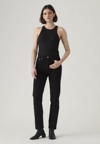 Black high-waisted jeans paired with a black ribbed tank top. Accessories include a button and subtle stitching details. Footwear features chunky heels.