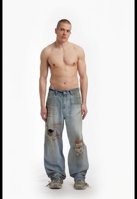 Jeans de denim azul claro com um corte relaxado, apresentando grandes rasgos e desbotamento. Combinados com sapatilhas bejes, o modelo está posicionado contra um fundo branco.
