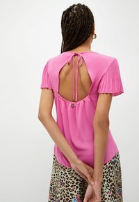 Blusa rosa con maniche corte plissettate, caratterizzata da un'apertura a forma rotonda sul retro e chiusura con fiocco. Tessuto morbido con una texture liscia.