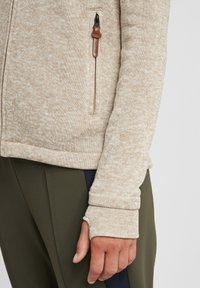Light beige zip-up jacket with a textured knit. Features a side pocket, thumbhole cuffs, and snug fit at the hem.