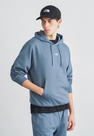 Noor mees, kes kannab sinist The North Face kapuutsiga tunked ja sobivaid pükse, musta mütsi, vaadates küljele, üks käsi kapuutsitaskus, neutraalne taust.