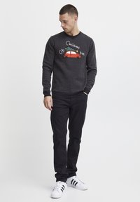Maglione nero con pois bianchi, caratterizzato da una grafica colorata di un'auto rossa che trasporta un albero di Natale, indossato con pantaloni neri e sneakers bianche.