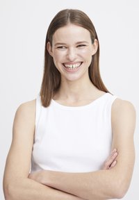 Model trägt ein weißes, geripptes Tanktop mit rundem Ausschnitt, lächelt, die Haare sind glatt und fallen bis zu den Schultern, Arme sind verschränkt.