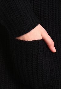 Gros plan d'un pull en tricot noir, présentant une texture côtelée, une poche visible et un design de poignet ajusté. Matière douce avec des points bien définis.