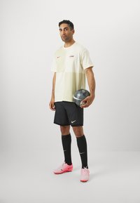Tenue de sport pour homme : t-shirt à blocs de couleur crème, short noir, chaussures de football roses, chaussettes noires, tenant un ballon de football gris. Logo Nike visible.