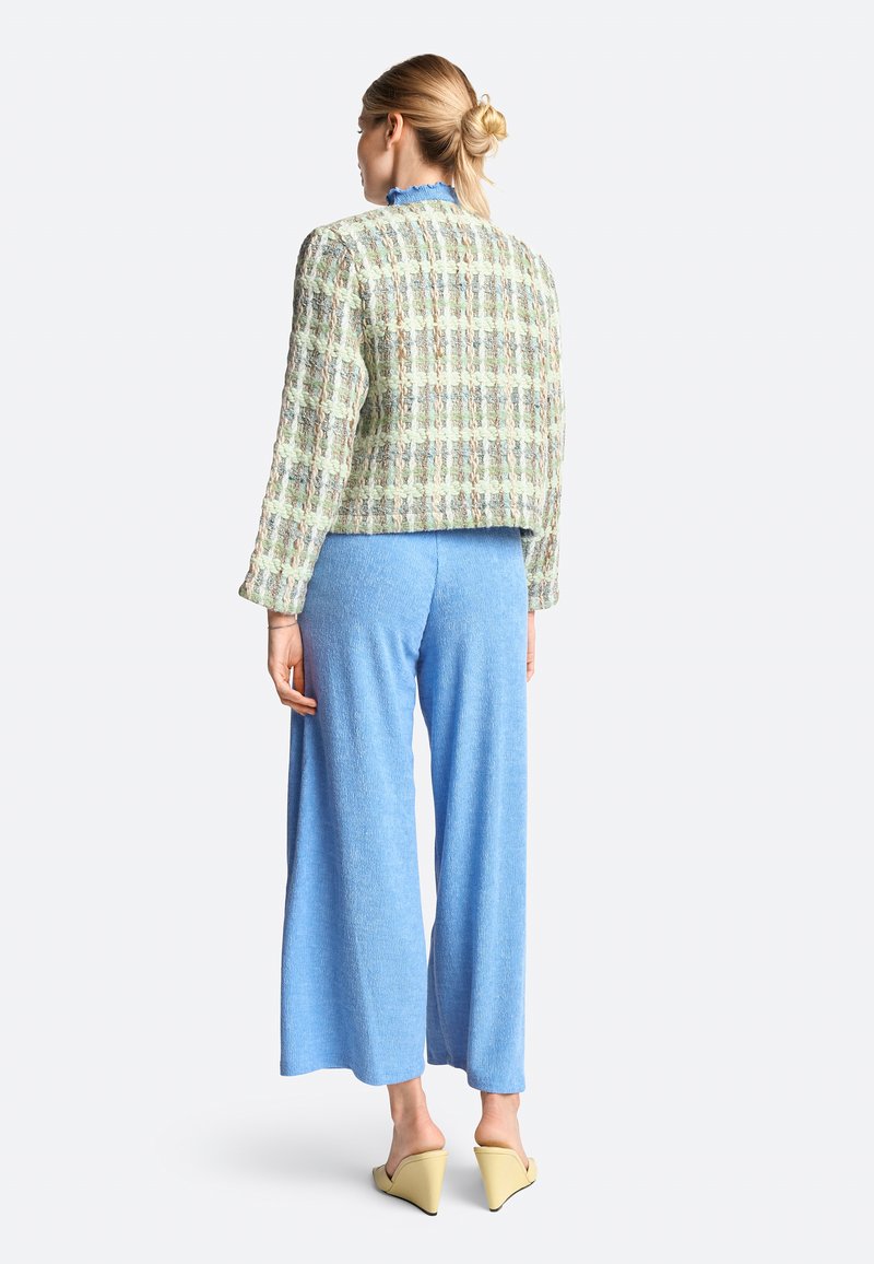 Hellgrüne, strukturierte Tweedjacke mit einem cropped Design, kombiniert mit blauen weiten Hosen und gelben Mules. Rückansicht, mit Rüschendetail am Kragen.