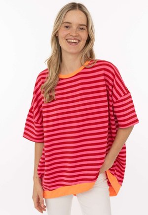 Lächelnde Frau mit langen blonden Haaren, trägt ein lockeres rot-pink gestreiftes Hemd mit orangem Besatz und weiße Hose, posiert mit der Hand in der Tasche.
