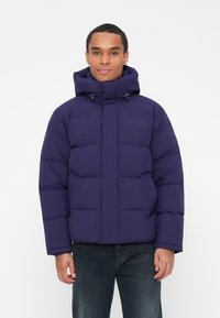 Man wearing a dark purple padded winter jacket with a high collar and hood, paired with dark blue jeans, standing against a white background.