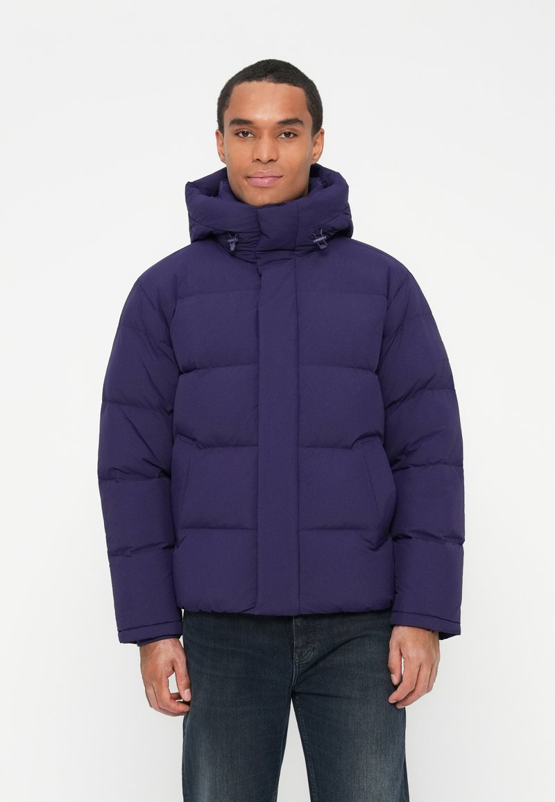 Man wearing a dark purple padded winter jacket with a high collar and hood, paired with dark blue jeans, standing against a white background.