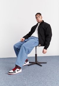 Black bomber jacket, white crew neck shirt, loose blue jeans, and black, red, and white sneakers. Seated on a black metal stool.