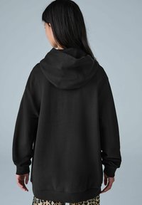 Sudadera negra holgada con una capucha amplia, mangas largas y puños de canalé. La tela parece suave y tiene detalles de costura mínimos.