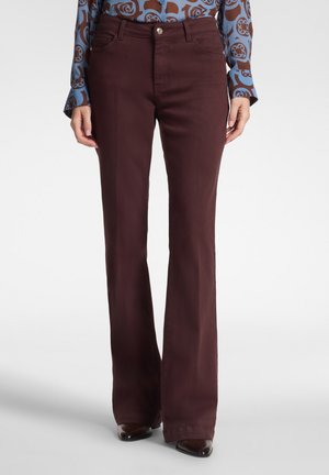 Donna che indossa pantaloni a zampa marrone scuro e una camicia blu a maniche lunghe con fantasia, in piedi su sfondo bianco.