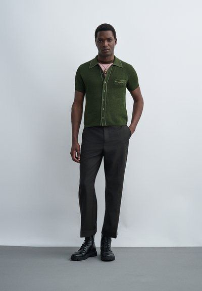 Νεαρός άνδρας στέκεται με το ένα χέρι στην τσέπη, φοράει σκούρο πράσινο κοντομάνικο πλεκτό πουκάμισο, μαύρο παντελόνι, μαύρα μποτάκια με κορδόνια και στρωμένα κολιέ.