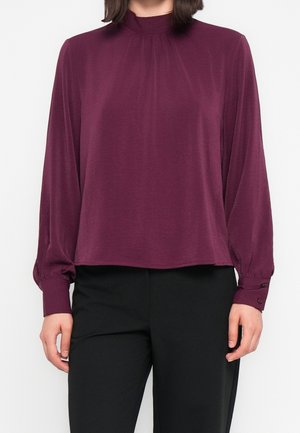 Femme portant un chemisier prune à manches longues et col montant, avec des détails froncés et des poignets boutonnés, associé à un pantalon noir tailleur.