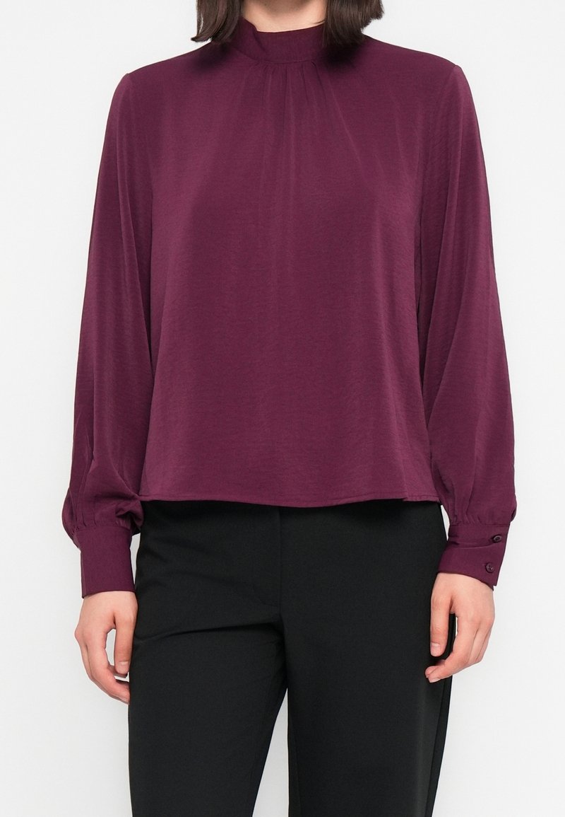Femme portant un chemisier prune à manches longues et col montant, avec des détails froncés et des poignets boutonnés, associé à un pantalon noir tailleur.