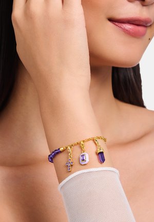 Armband in Goldton mit lila Akzentperlen und drei Anhängern: ein Kreuz, ein facettierter Stein und ein spitzer Kristall. Strukturierte Mesh-Hülle sichtbar.