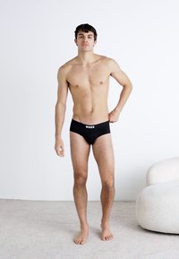 Culotte noire en coton avec une ceinture logo, design ajusté et texture lisse. Le modèle se tient devant un fond uni sur un tapis.