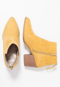 Un par de botas de ante amarillo mostaza hasta el tobillo con tacones de bloque de madera apilados y cremalleras laterales sobre un fondo blanco.