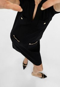 Svart klänning med korta ärmar, två bröstfickor och gulddetaljer. Har gulddragkedjor på sidfickorna och en figurnära silhuett. Svarta spetsiga skor.