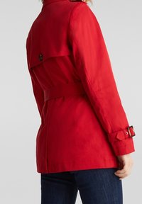 Abrigo rojo estilo trench con cintura ceñida, abertura trasera y detalles de botones. Tejido suave con mangas largas y tirantes ajustables en los puños.