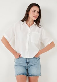 White short-sleeve shirt made of lightweight fabric, featuring a collar, a single front pocket, and a relaxed fit, paired with denim shorts.