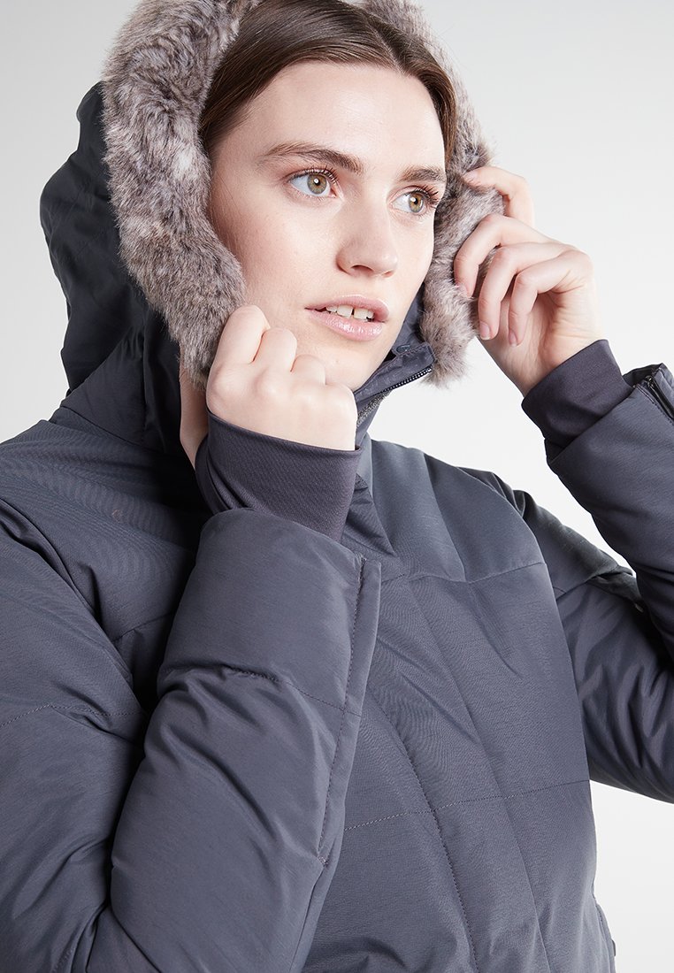 Mujer ajustando la capucha de una chaqueta de invierno gris oscuro con borde de piel, mirando hacia un lado contra un fondo claro y liso.