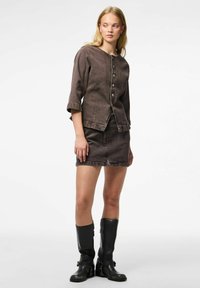 Giubbotto di jeans marrone con maniche a tre quarti e bottoni frontali, abbinato a una gonna di jeans marrone coordinata. Stivali neri al ginocchio completano l'outfit.
