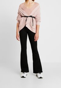 Pull rose clair en maille torsadée oversize avec un col en V, cintré par une ceinture noire, associé à un pantalon noir évasé et des baskets blanches.