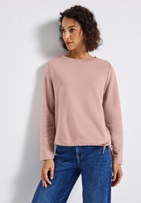Ljusrosa sweatshirt med rund halsringning, långa ärmar och knytdetalj på sidan. Slät textur, avslappnad passform, i kombination med blå jeans.