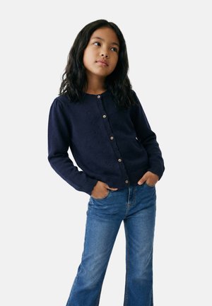 Fille aux longs cheveux foncés portant un cardigan bleu marine boutonné et un jean bleu, debout les mains dans les poches, regardant légèrement sur le côté.