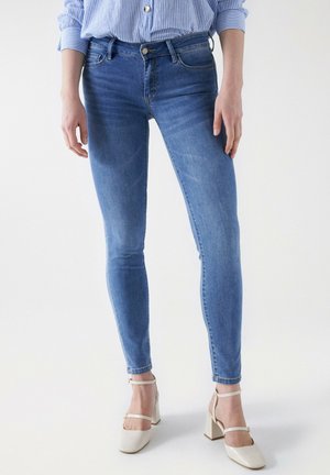 Jeans ajustados en denim azul de lavado medio, con diseño de cinco bolsillos, presillas para el cinturón y una tapeta de botones. Combinados con zapatos de tacón cuadrado en color blanco roto.