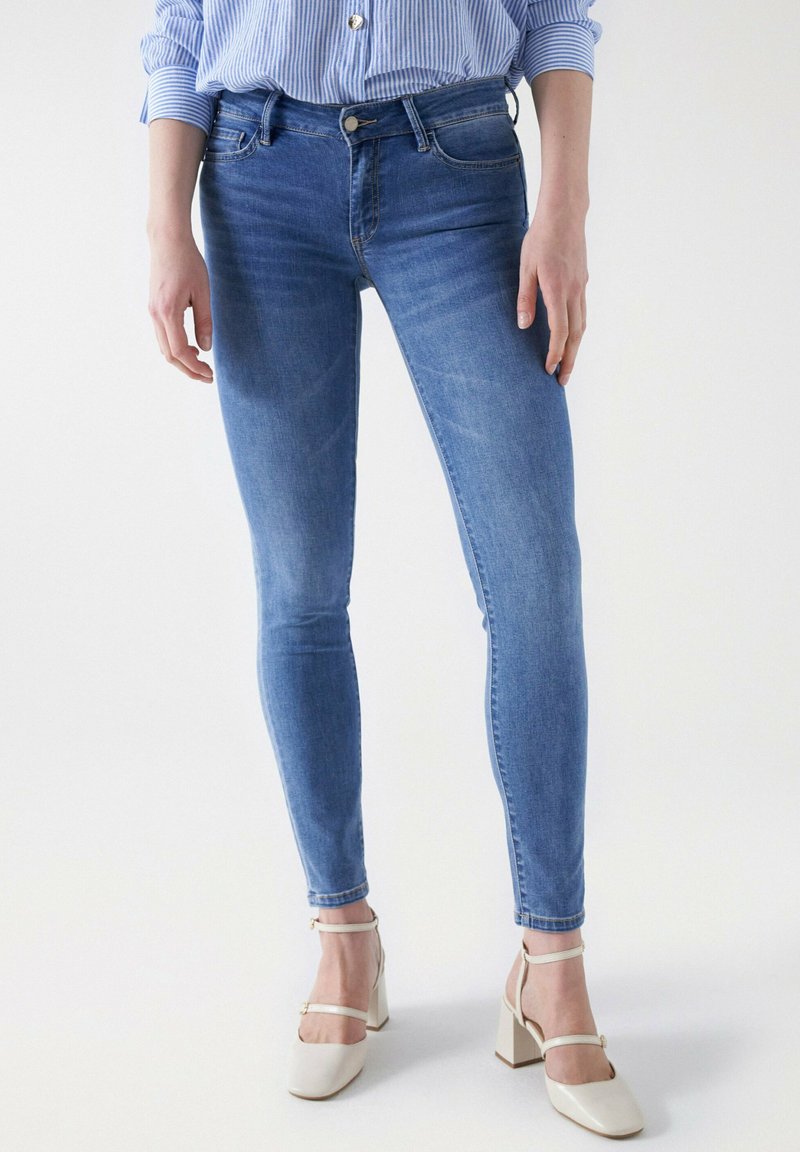Schlanke Jeans aus mittelblauem Denim mit einem Fünf-Taschen-Design, Gürtelschlaufen und einem Knopfverschluss. Getragen mit off-white Blockabsatzschuhen.