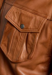 Nahaufnahme einer braunen Lederjacke mit einer großen Klappentasche auf der Brust, die mit einem silbernen Druckknopf verschlossen ist. Sichtbare Nähte und eine glatte Textur.