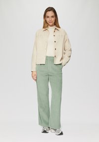 Beige corduroy jas met houten knopen, witte geribde trui en lichtgroene flanellen wijde broek. Zilveren sneakers maken de outfit compleet.