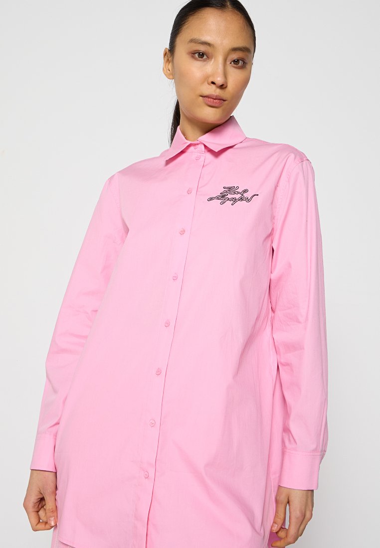 Chemise à manches longues en coton rose clair avec un col pointu et un texte brodé noir sur le côté gauche. Texture lisse, coupe décontractée.