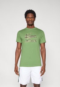 Πράσινο βαμβακερό t-shirt με μαύρο και μπεζ γραφικό λογότυπο. Το πουκάμισο έχει στρογγυλή λαιμόκοψη και κοντά μανίκια, σε συνδυασμό με λευκές βερμούδες.