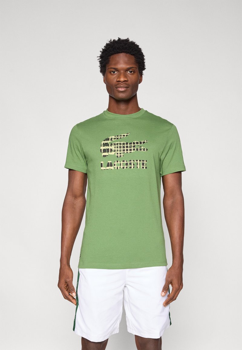 Πράσινο βαμβακερό t-shirt με μαύρο και μπεζ γραφικό λογότυπο. Το πουκάμισο έχει στρογγυλή λαιμόκοψη και κοντά μανίκια, σε συνδυασμό με λευκές βερμούδες.