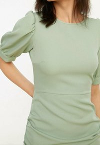 Robe vert clair avec des manches courtes bouffantes, un corsage ajusté et une texture lisse. Présente un col rond simple et un léger ruché à la taille.