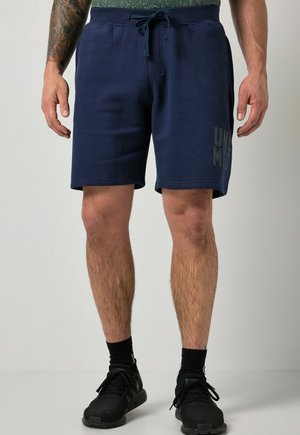 Mann trägt marineblaue Shorts mit Kordelzug, schwarze Socken und schwarze Sportschuhe, steht vor einem schlichten Hintergrund.