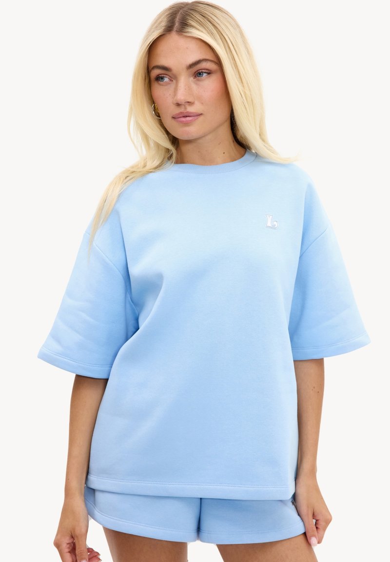 Femme blonde portant un sweat-shirt ample bleu clair à manches courtes et un short assorti, debout devant un fond uni.