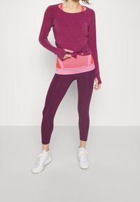 Top met lange mouwen in bessenrood met een knoopdetail, gedragen over een roze-oranje aansluitende tanktop en bordeauxrode leggings. Witte sportschoenen maken de look compleet.