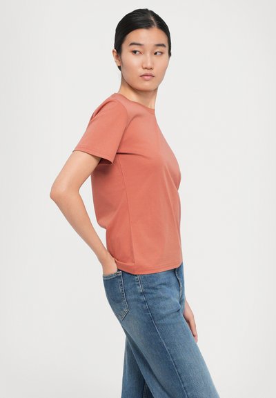 Ung kvinna med mörkt hår i en låg knut, klädd i en rostfärgad t-shirt med kort ärm och blå jeans, som tittar åt sidan med en hand i fickan.